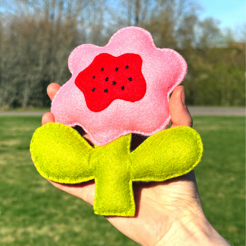 Hand holding a pink flower-shaped toy with a red center and green leaves outdoors.