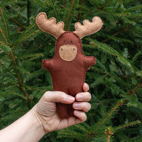 Hand holding a brown moose felt toy against a green tree background