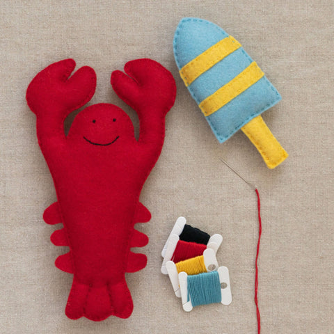 Red lobster plush toy and blue and yellow striped buoy and threads on a neutral fabric background