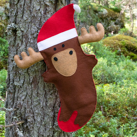 Moose-shaped Christmas stocking with a Santa hat attached to a tree in a forest setting.