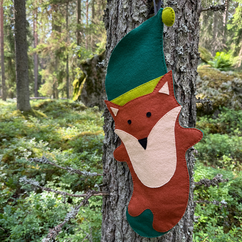 Felt fox ornament with green hat hanging on a tree in a forest setting