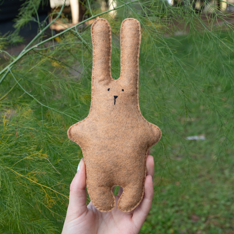 Brown felt bunny toy held in a hand with green foliage in the background