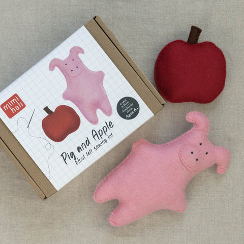 Pink felt pig and red apple toy set with packaging on a beige surface