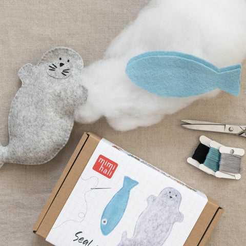 Crafting materials including a felt seal, fish, and a packaging with sewing supplies on a beige surface.